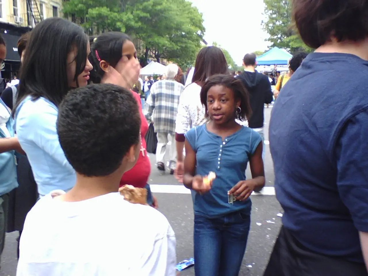 In this image there are group of persons standing and walking. In the background there are trees,...