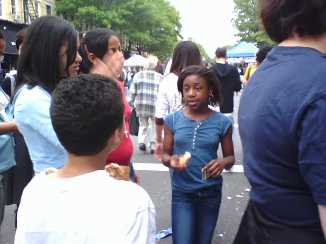 In this image there are group of persons standing and walking. In the background there are trees,...