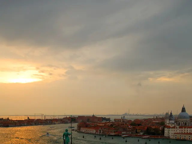 This picture shows few buildings and we see few boats in the water and a cloudy sky with sunlight.